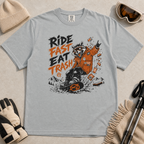 Granite gray t-shirt with a graphic of a raccoon in an orange jacket and text 'Ride Fast Eat Trash' on a beige surface.