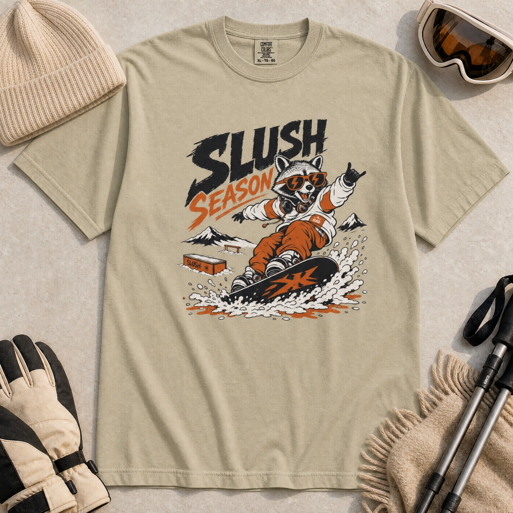 Khaki colored t-shirt with a graphic of a raccoon snowboarding and 'Slush Season' text, surrounded by winter accessories.