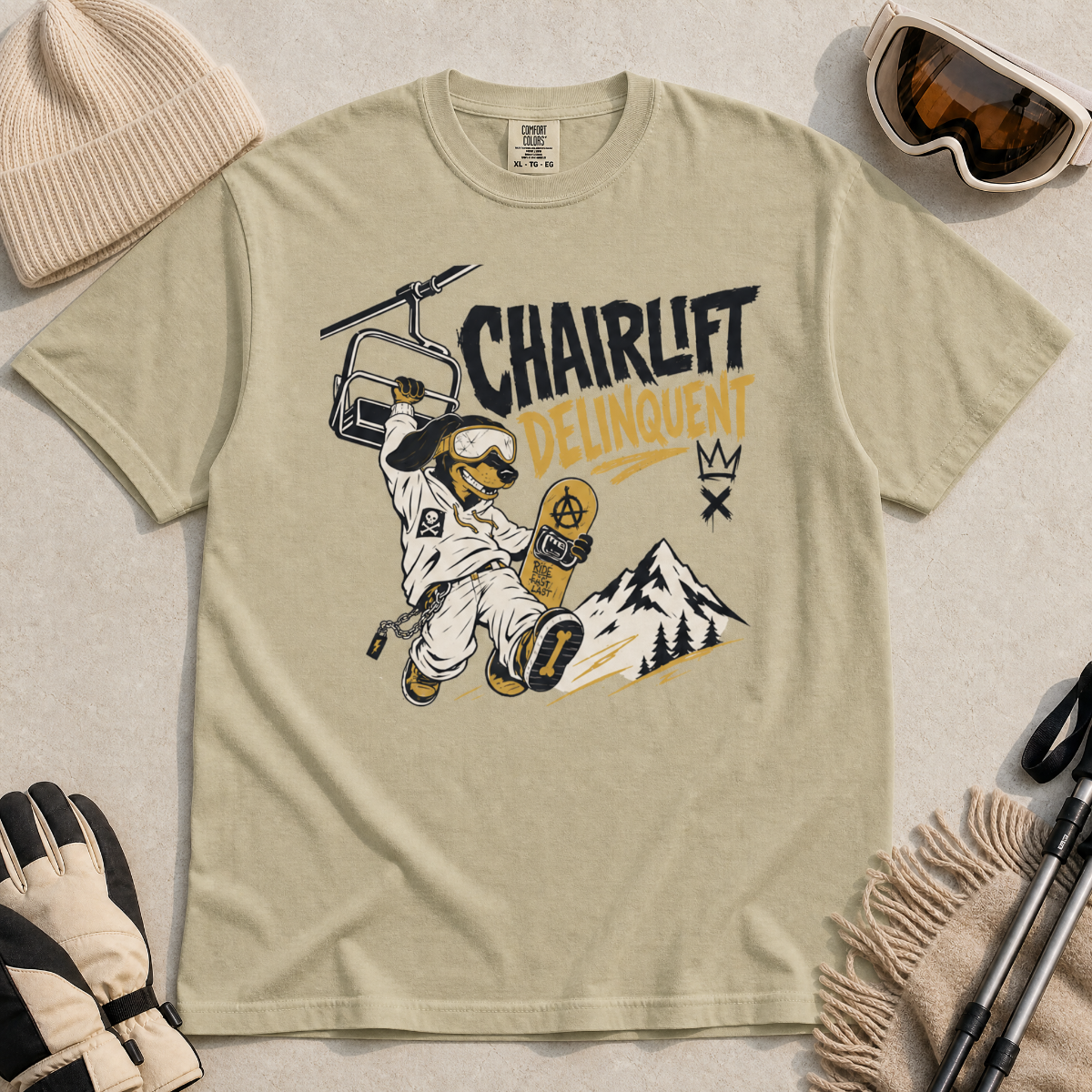 Bay green t-shirt with a graphic design featuring a dachshund snowboarder and 'Chairlift Delinquent' text, surrounded by winter accessories.