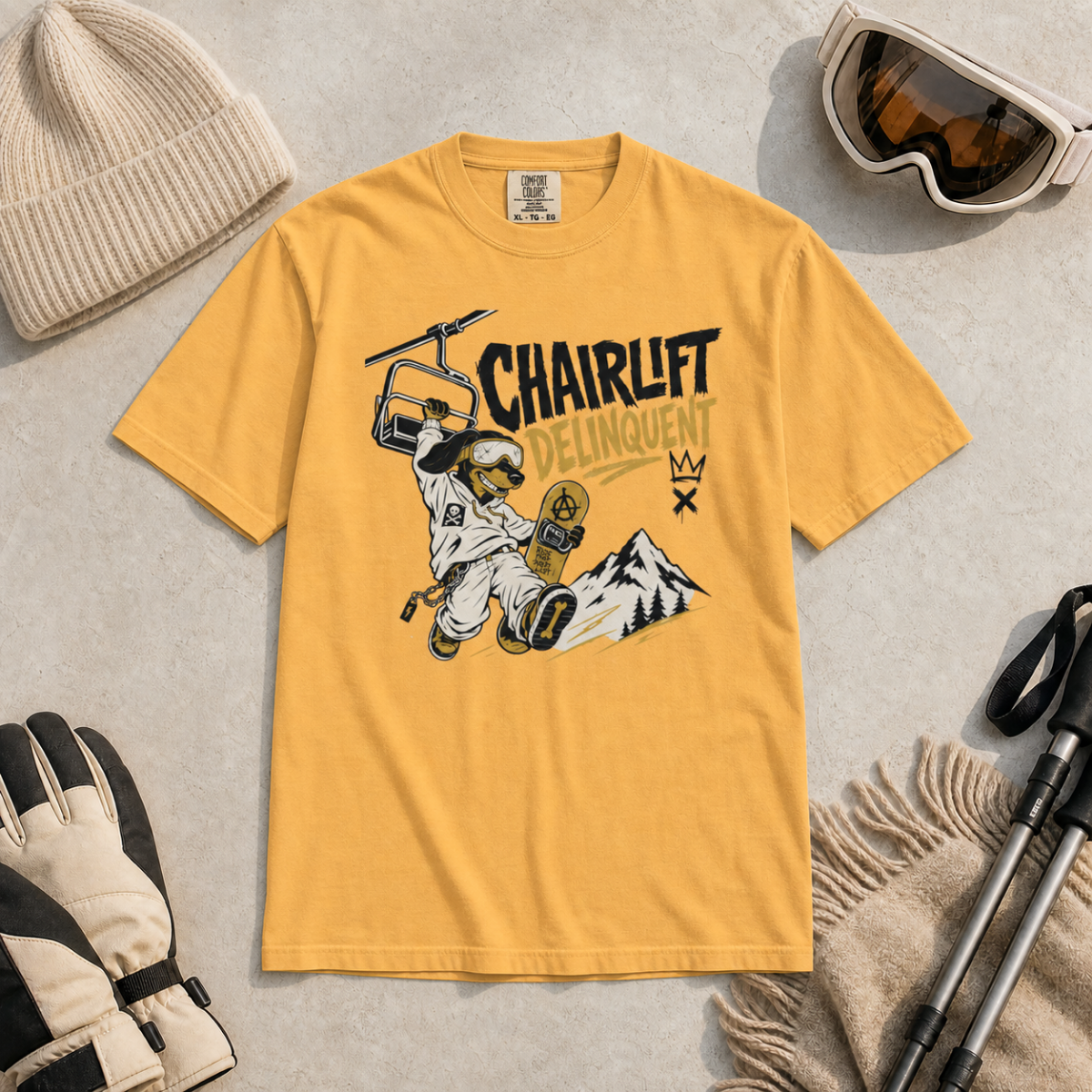 Mustard yellow t-shirt with a graphic design of a skier and chairlift, surrounded by winter gear on a light gray background.