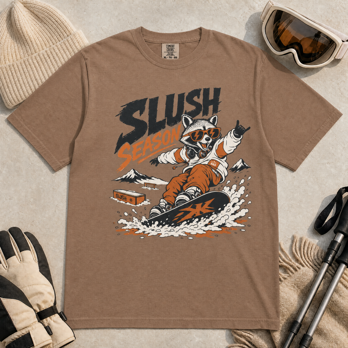 Espresso brown colored t-shirt with a graphic of a raccoon snowboarding and 'Slush Season' text, surrounded by winter accessories.