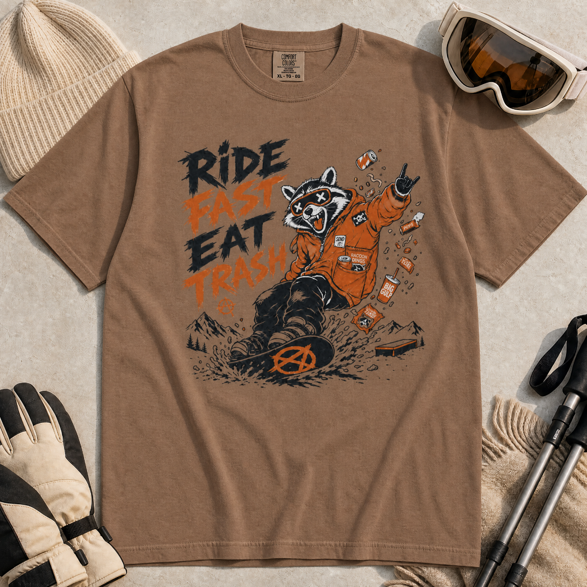 Espresso brown t-shirt with a graphic of a raccoon in an orange jacket and text 'Ride Fast Eat Trash' on a beige surface.