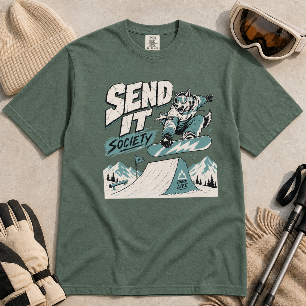 Blue spruce t-shirt with a graphic design of a snowboarding wolf and text 'Send It Society' on a beige surface with winter accessories.