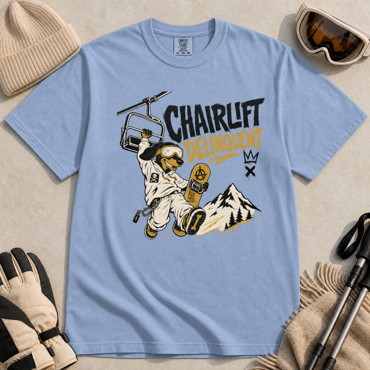 Washed denim blue t-shirt with a graphic design featuring a dachshund snowboarder and 'Chairlift Delinquent' text, surrounded by winter accessories.