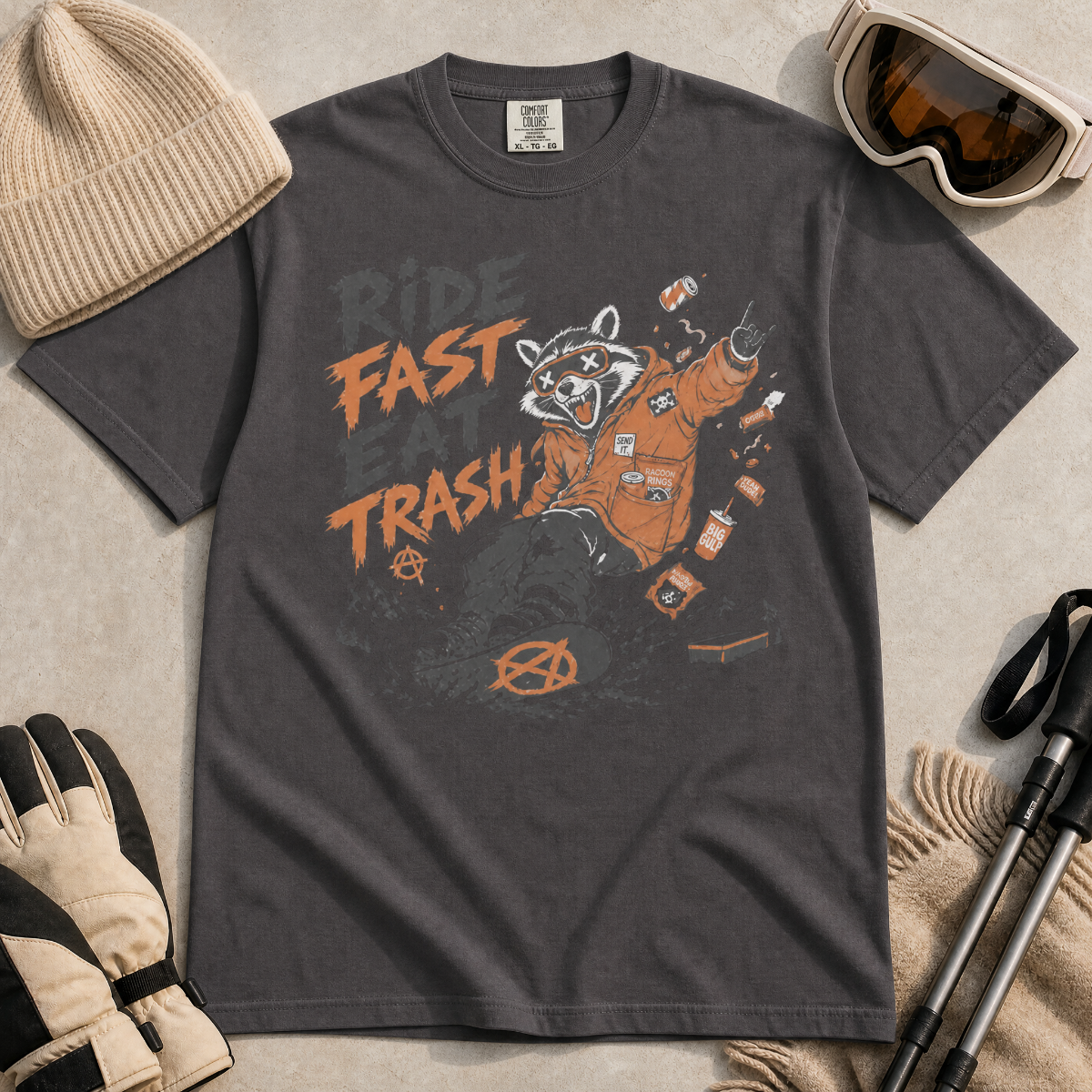 Graphite gray t-shirt with a graphic of a raccoon in an orange jacket and text 'Ride Fast Eat Trash' on a beige surface.