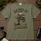 Moss green t-shirt with a graphic of an otter on a bike and text 'Send it for the snacks'.