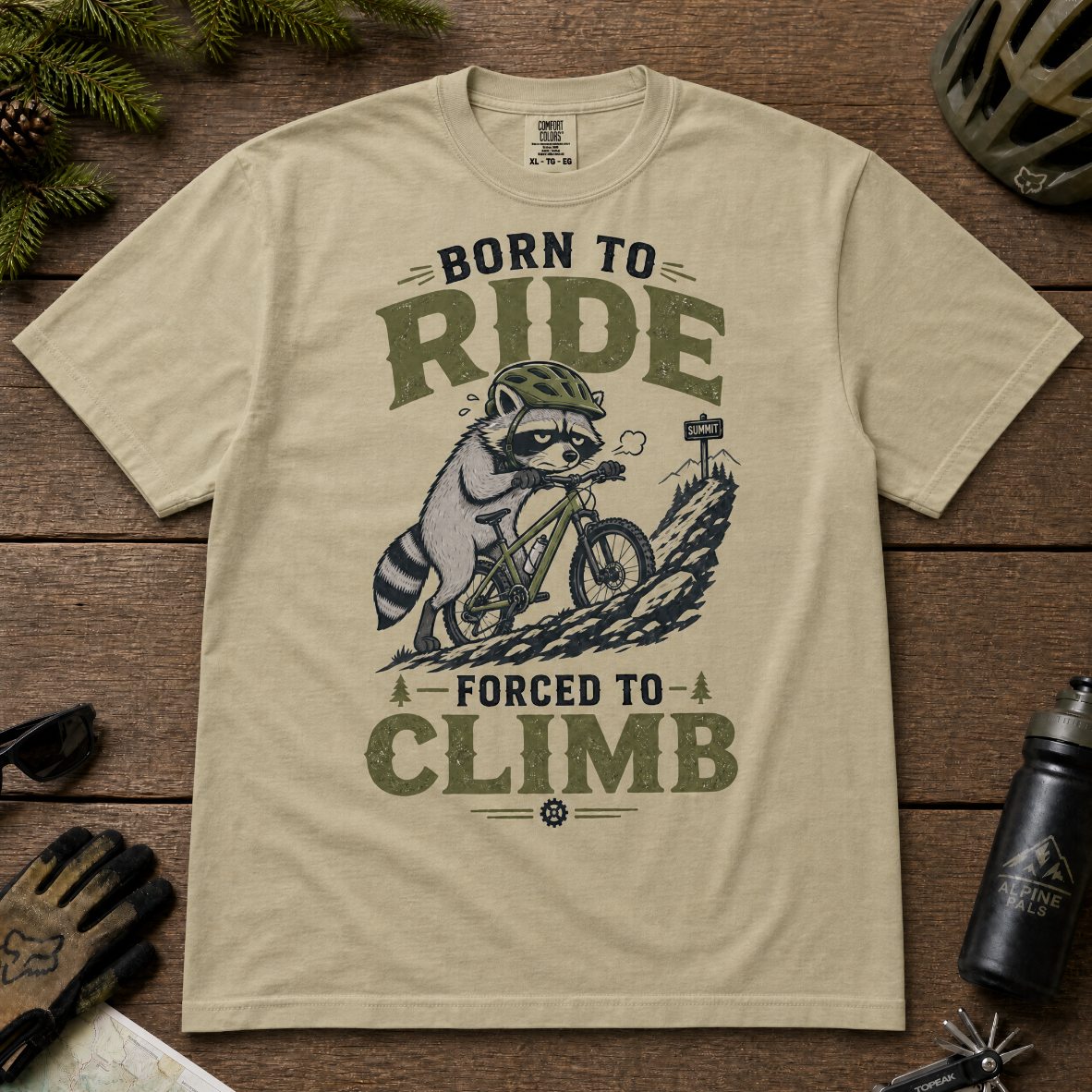 Khaki colored t-shirt with a graphic of a raccoon riding a bike and text 'Born to Ride, Forced to Climb'.