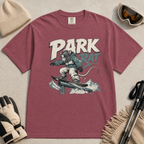 Brick red t-shirt with 'Park Rat' graphic design on a light gray surface with accessories.