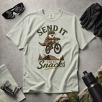 Bay green t-shirt with an otter riding a bike graphic and text 'Send it for the snacks'.