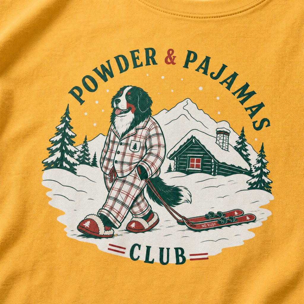 Mustard yellow t-shirt with a graphic of a dog in checkered pajamas pulling skis, trees, and a cabin, with 'Powder & Pajamas Club' text.