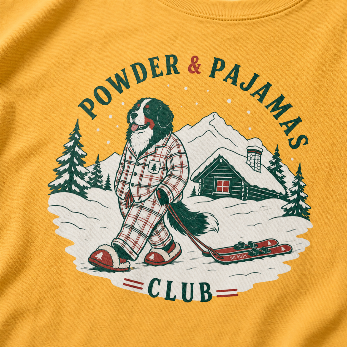 Mustard yellow t-shirt with a graphic of a dog in checkered pajamas pulling skis, trees, and a cabin, with 'Powder & Pajamas Club' text.