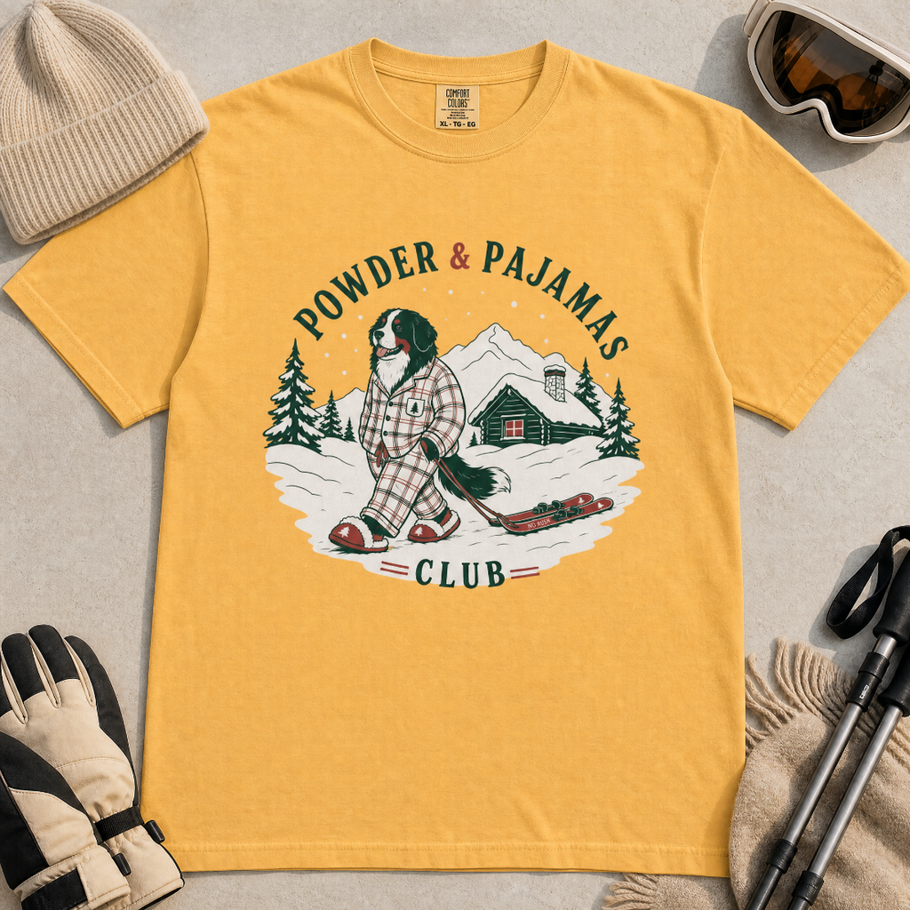 Mustard yellow t-shirt with a graphic of a dog in checkered pajamas pulling skis, trees, and a cabin, with 'Powder & Pajamas Club' text.