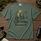 Blue spruce colored t-shirt with a graphic of a turtle riding a bike and text 'Slow Climbs, Fast Snacks'.