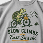 Granite colored t-shirt with a graphic of a turtle riding a bike and text 'Slow Climbs, Fast Snacks'.