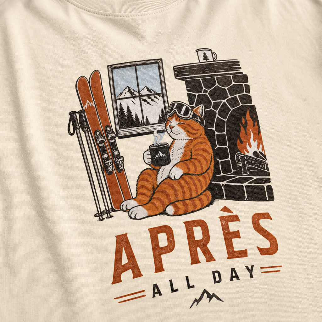 Ivory white t-shirt with a graphic of a cat by a fireplace and skis, with 'Après All Day' text.