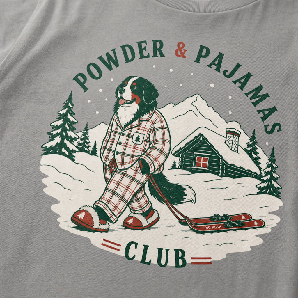 Grey t-shirt with a graphic of a dog in checkered pajamas pulling skis, trees, and a cabin, with 'Powder & Pajamas Club' text.