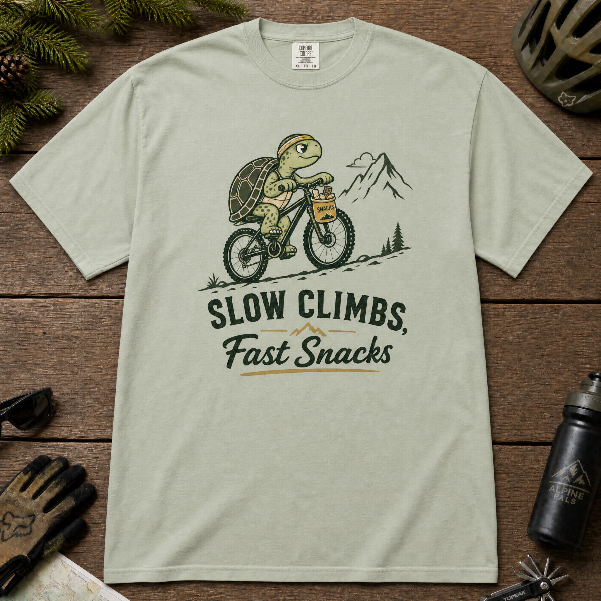 Bay green t-shirt with a graphic of a turtle riding a bike and text 'Slow Climbs, Fast Snacks'.