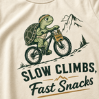 Ivory white t-shirt with a graphic of a turtle riding a bike and text 'Slow Climbs, Fast Snacks'.