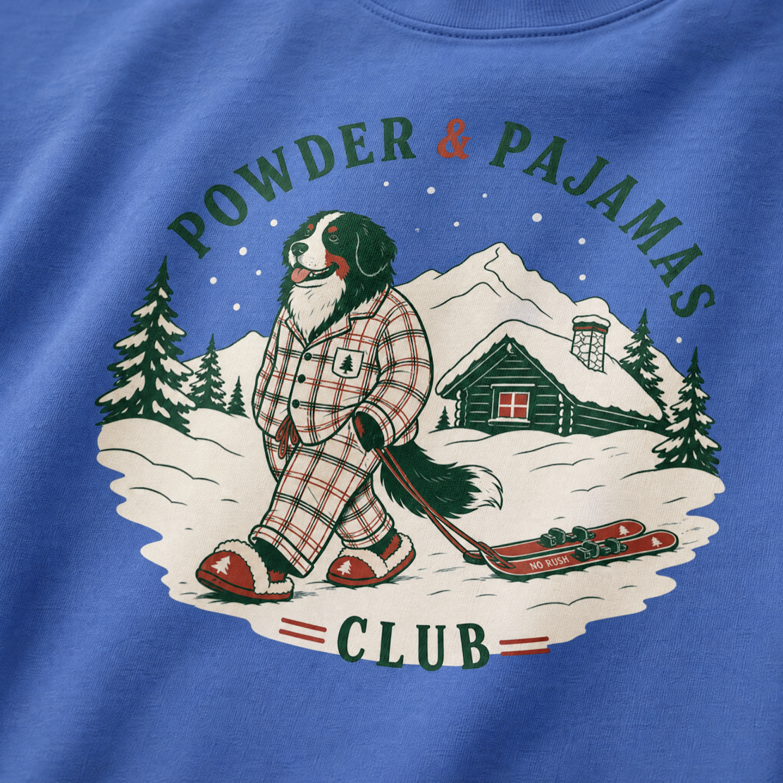 Mystic blue t-shirt with a graphic of a dog in checkered pajamas pulling skis, trees, and a cabin, with 'Powder & Pajamas Club' text.