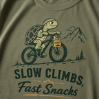 Moss green t-shirt with a graphic of a turtle riding a bike and text 'Slow Climbs, Fast Snacks'.
