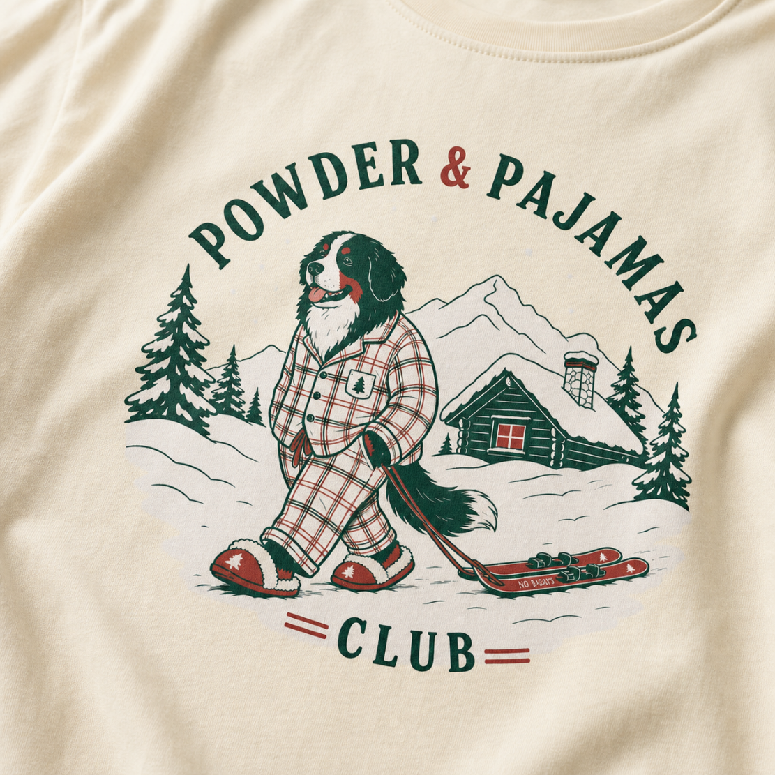 Ivory white t-shirt with a graphic of a dog in checkered pajamas pulling skis, trees, and a cabin, with 'Powder & Pajamas Club' text.
