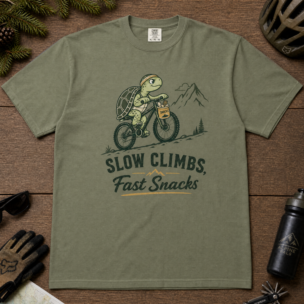 Moss green t-shirt with a graphic of a turtle riding a bike and text 'Slow Climbs, Fast Snacks'.