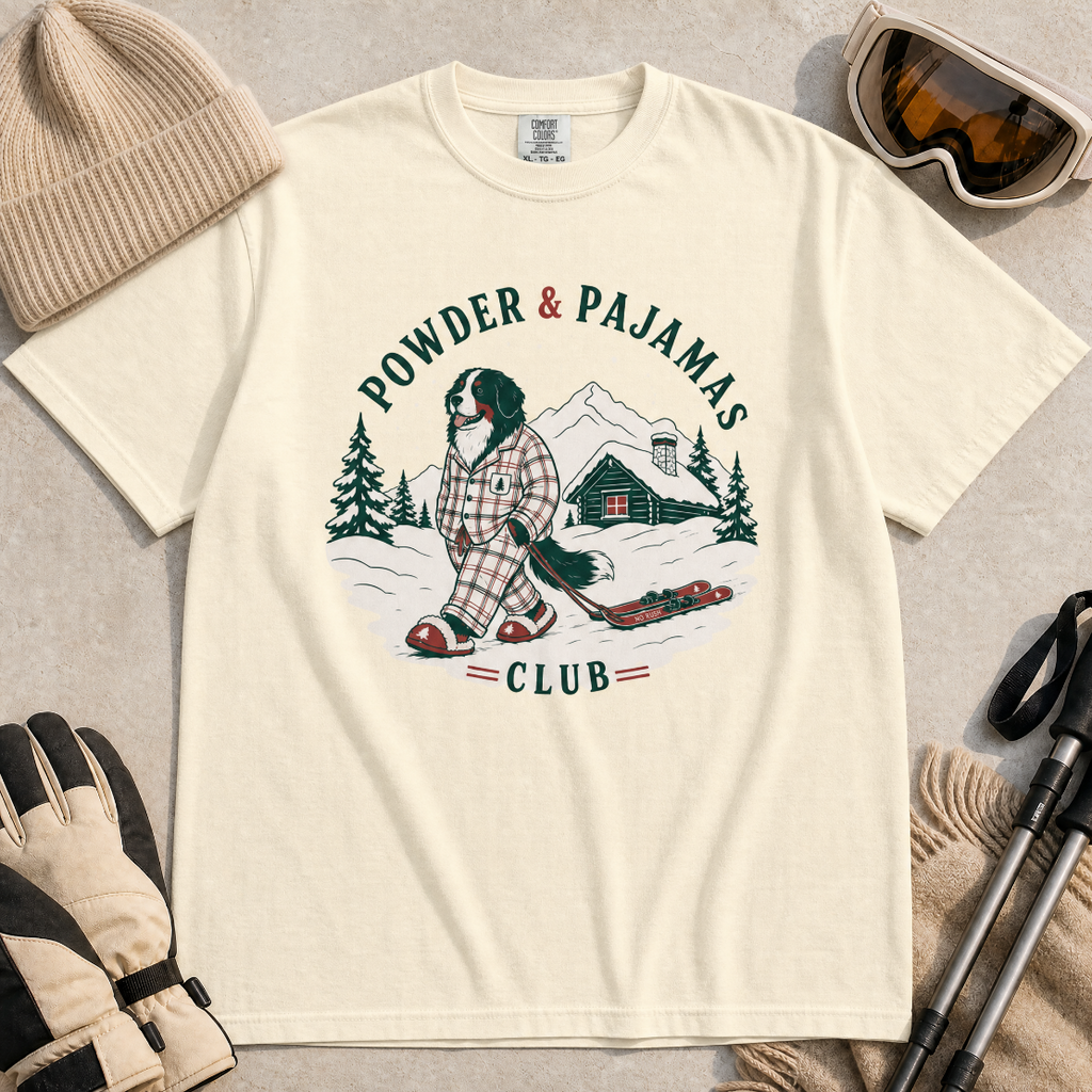 Ivory white t-shirt with a graphic of a dog in checkered pajamas pulling skis, trees, and a cabin, with 'Powder & Pajamas Club' text.