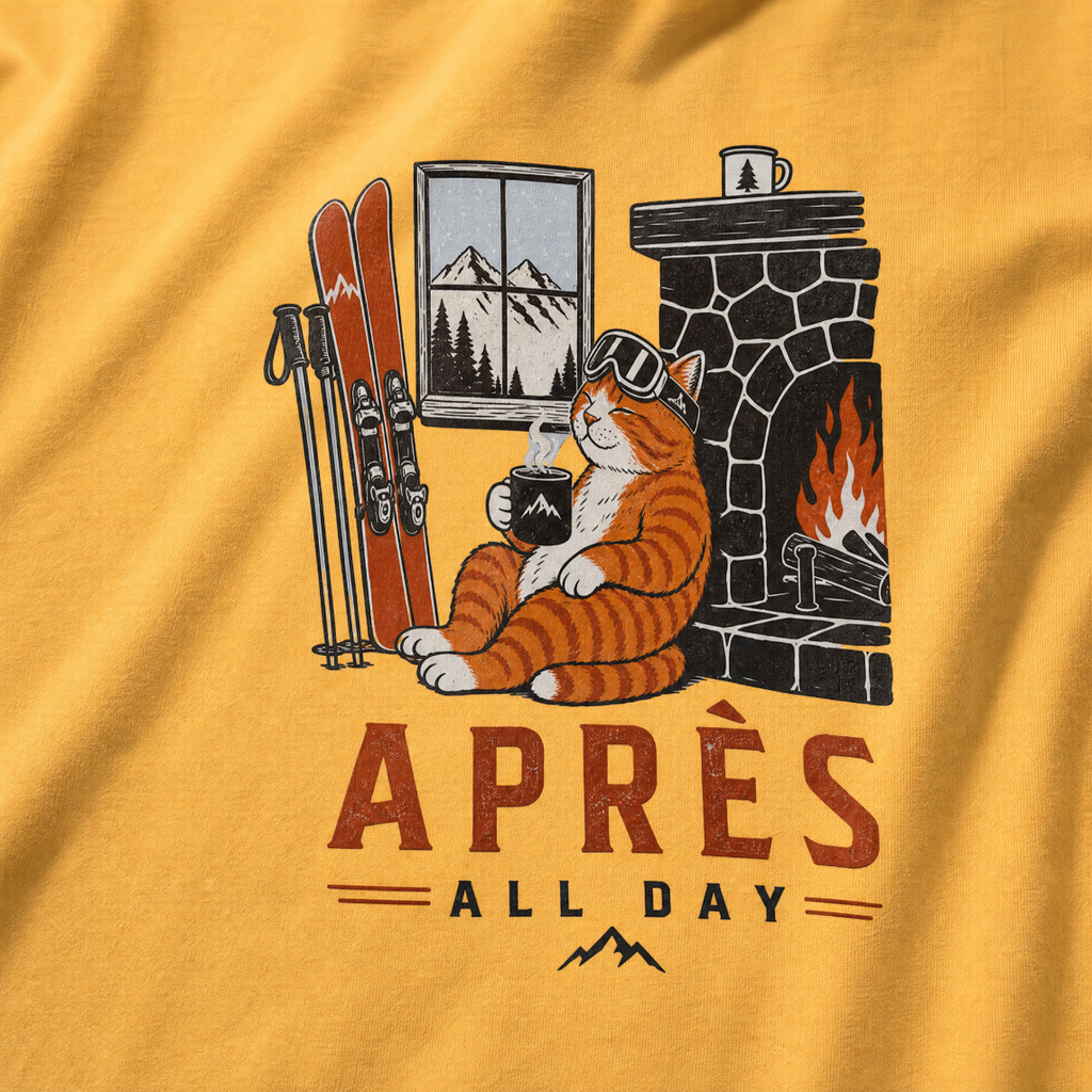 Mustard yellow t-shirt with a graphic of a cat by a fireplace and skis, with 'Après All Day' text.