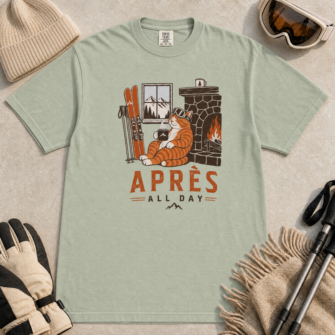 Bay green t-shirt with a graphic of a cat by a fireplace and skis, with 'Après All Day' text.