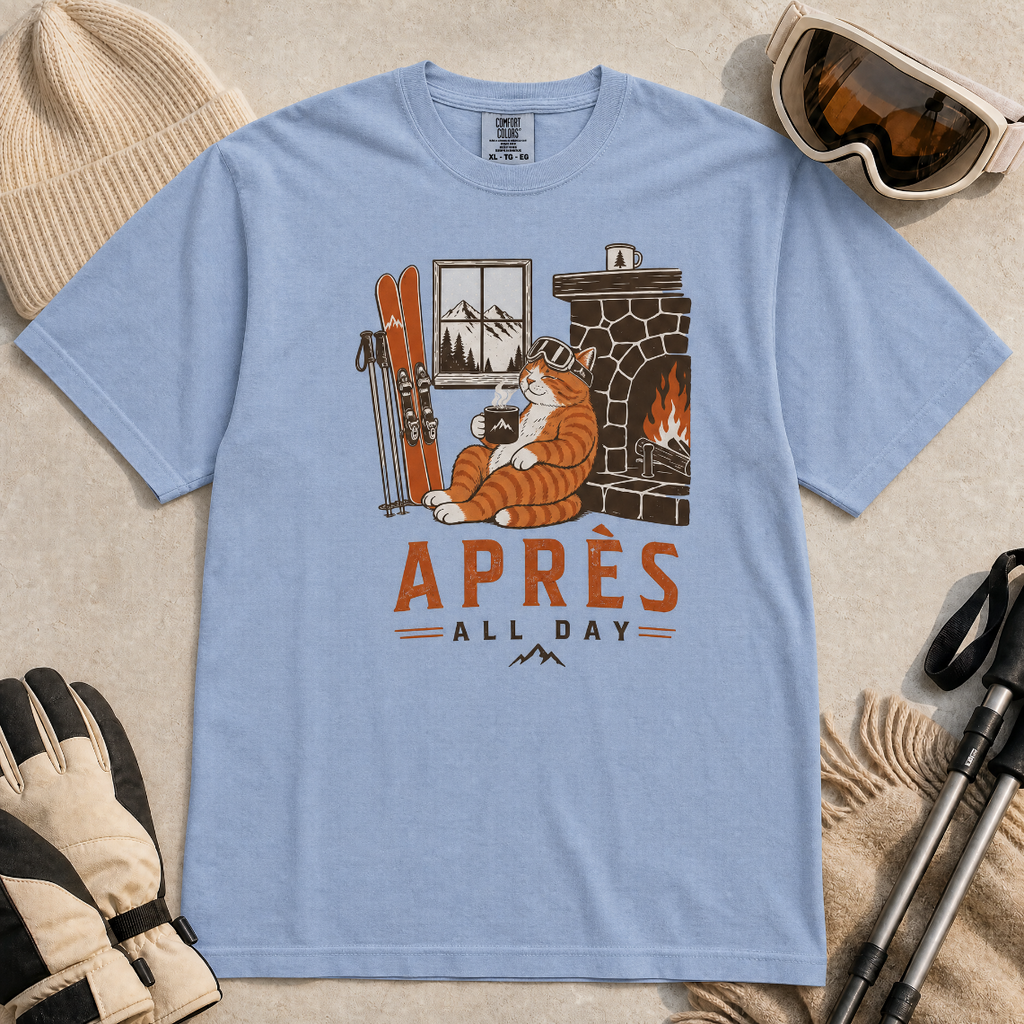 Washed denim blue t-shirt with a graphic of a cat by a fireplace and skis, with 'Après All Day' text.
