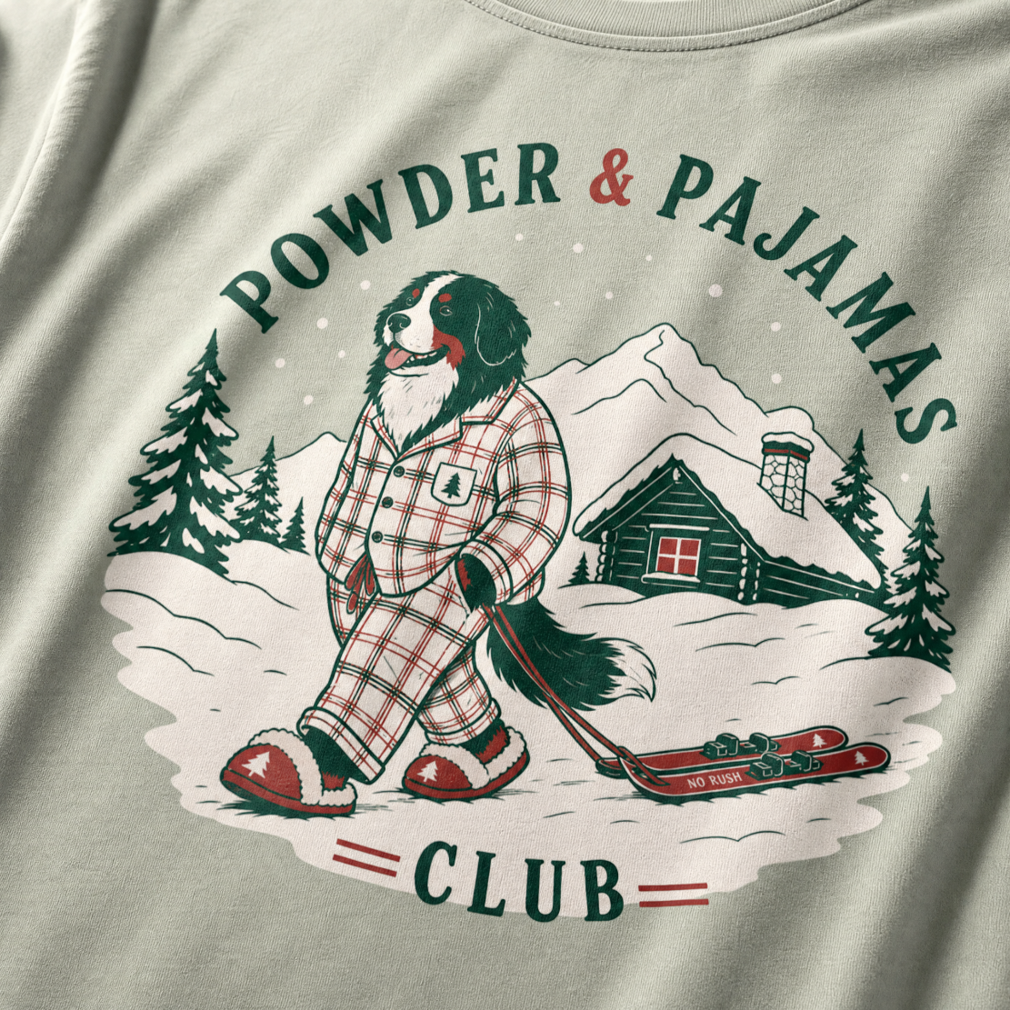 Bay green t-shirt with a graphic of a dog in checkered pajamas pulling skis, trees, and a cabin, with 'Powder & Pajamas Club' text.