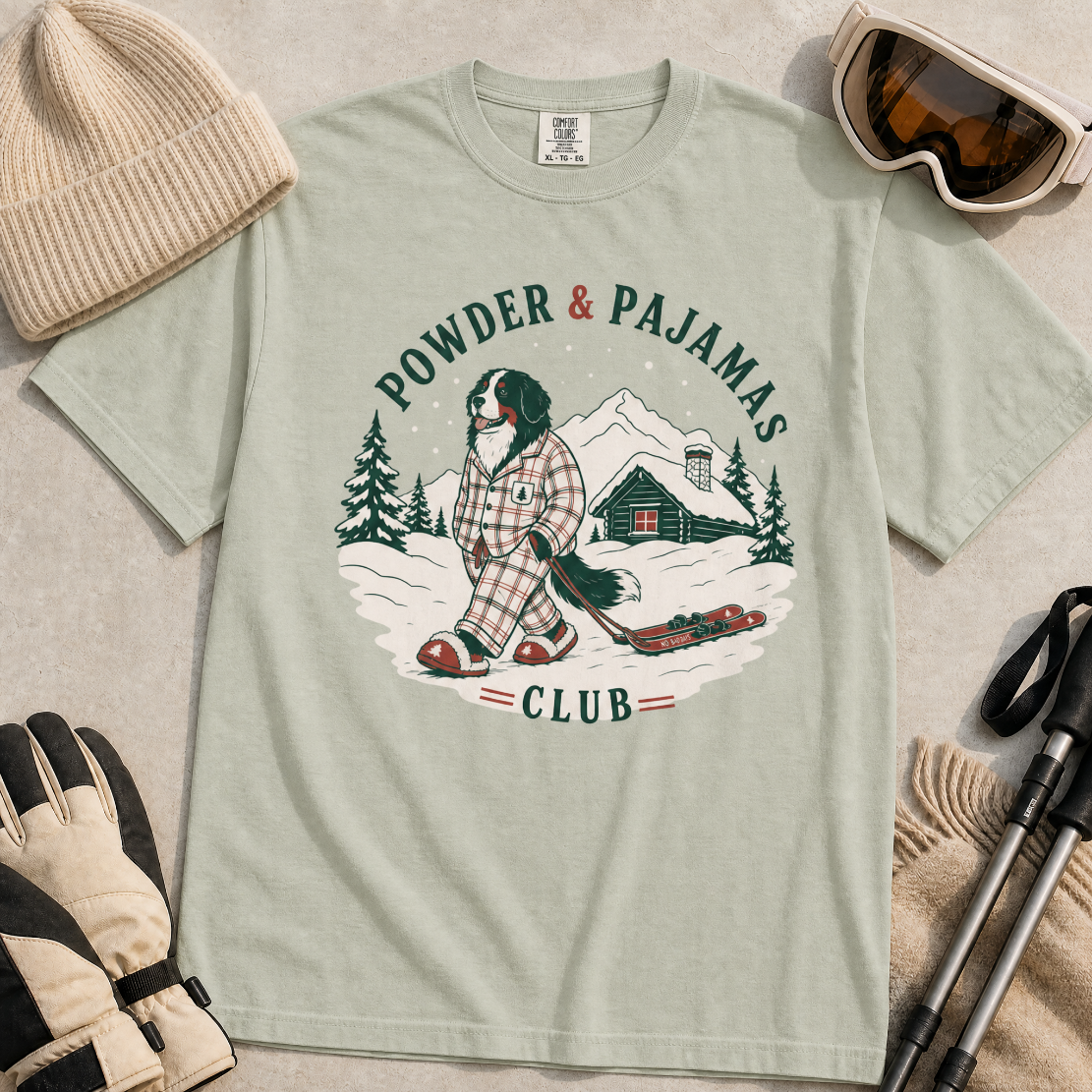 Bay green t-shirt with a graphic of a dog in checkered pajamas pulling skis, trees, and a cabin, with 'Powder & Pajamas Club' text.