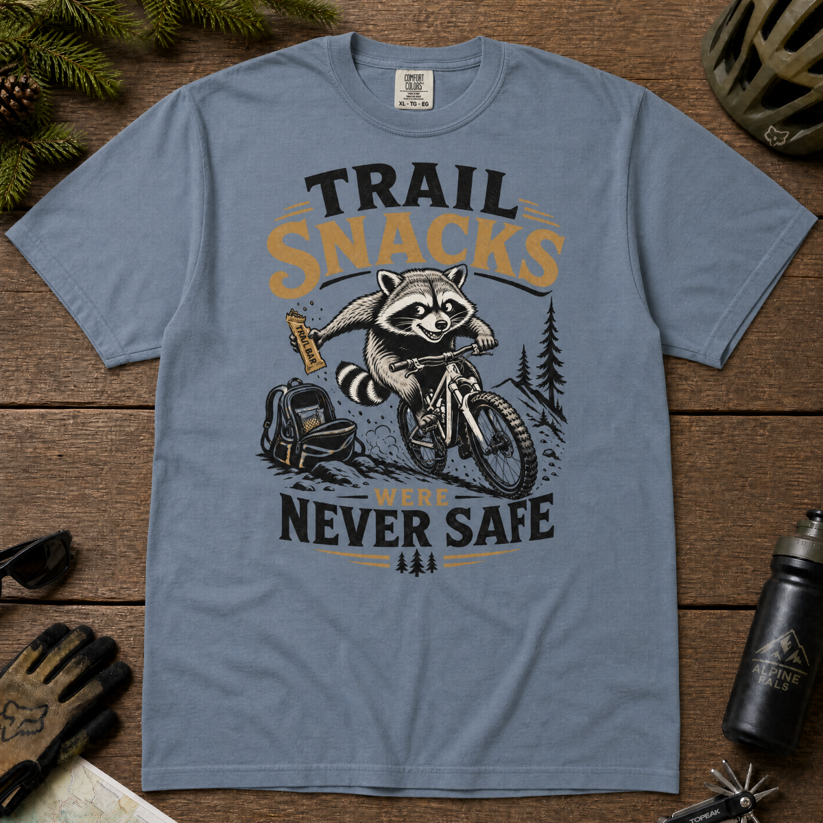 Blue jean t-shirt with a raccoon graphic and text 'Trail Snacks Were Never Safe' on a wooden surface.