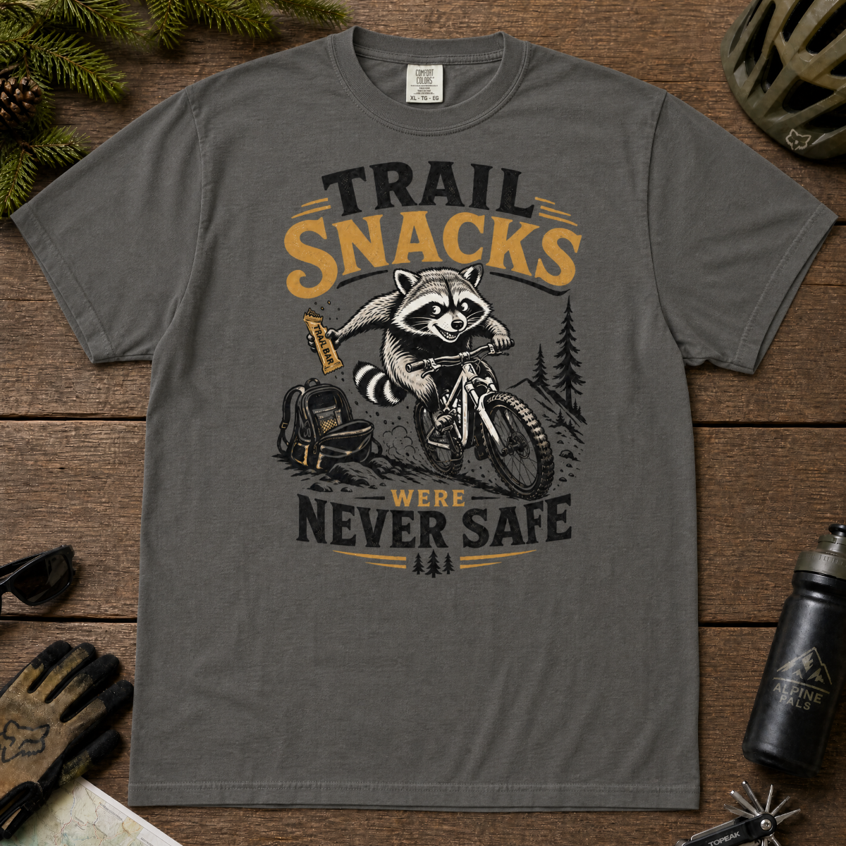 Pepper gray t-shirt with a raccoon graphic and text 'Trail Snacks Were Never Safe' on a wooden surface.