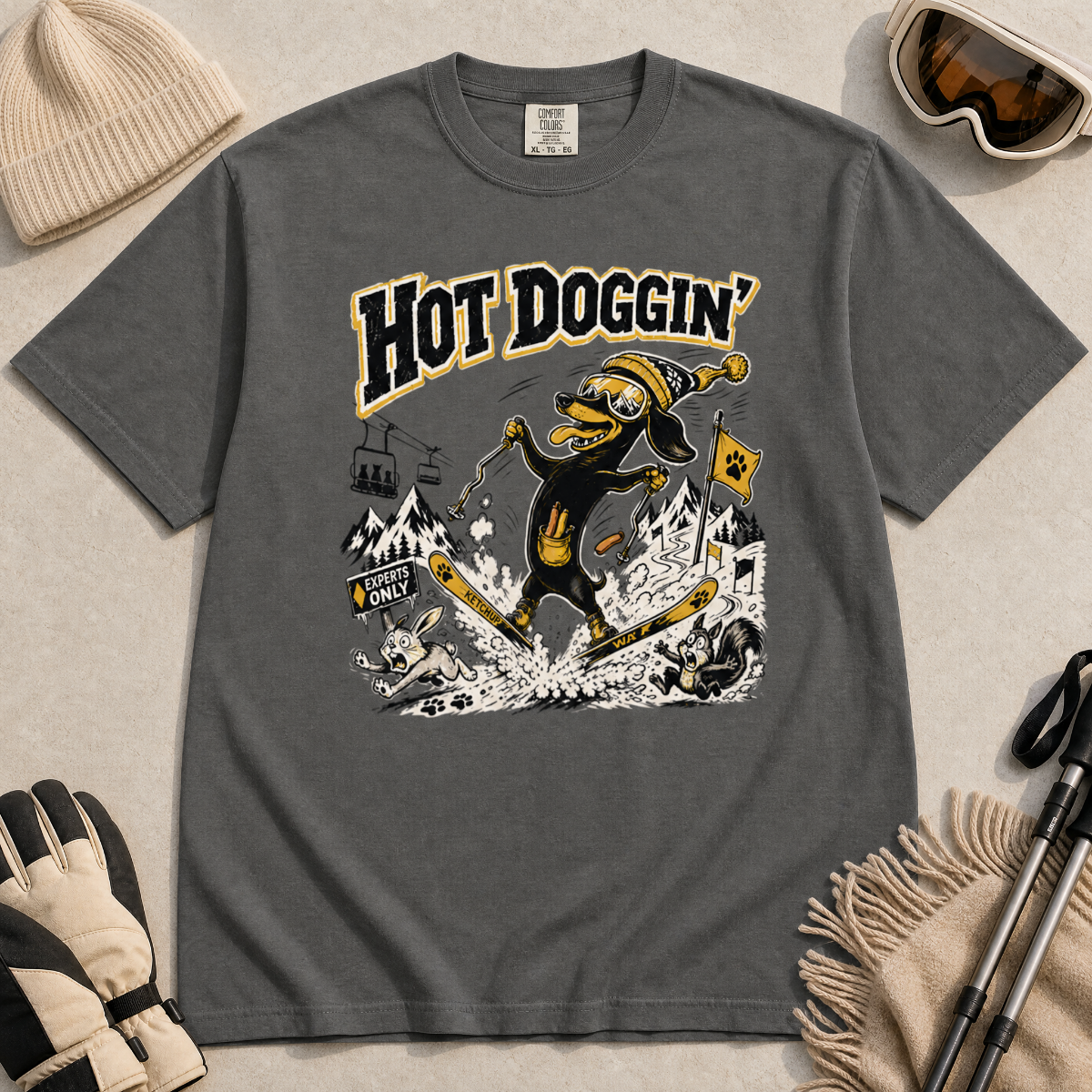 Pepper gray t-shirt with a graphic design of a dog skiing, text 'Hot Doggin', surrounded by winter accessories on a beige surface.