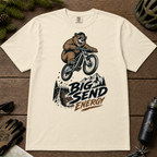 Ivory white t-shirt with bear riding a bike graphic and 'Big Send Energy' text on a wooden surface.