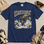 Navy blue t-shirt with a graphic design of a dog skiing, text 'Hot Doggin', surrounded by winter accessories on a beige surface.