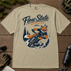 Khaki colored t-shirt with a fox riding a bike graphic and 'Flow State Activated' text on a wooden surface.