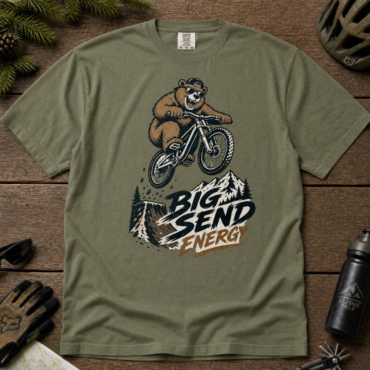 Moss green t-shirt with bear riding a bike graphic and 'Big Send Energy' text on a wooden surface.