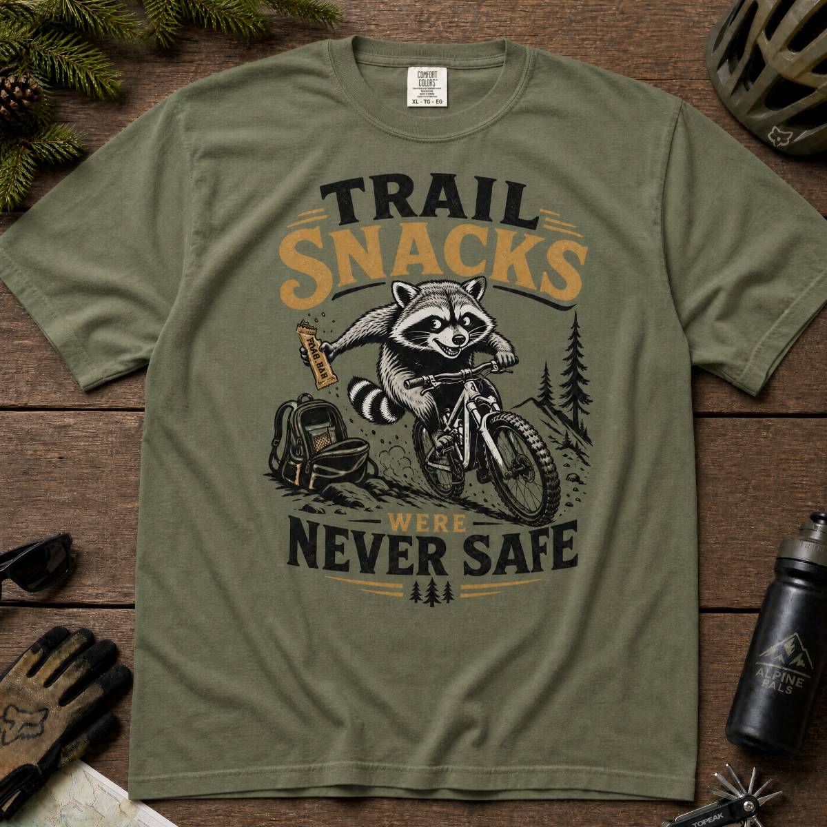 Moss green t-shirt with a raccoon graphic and text on a wooden surface with outdoor gear.
