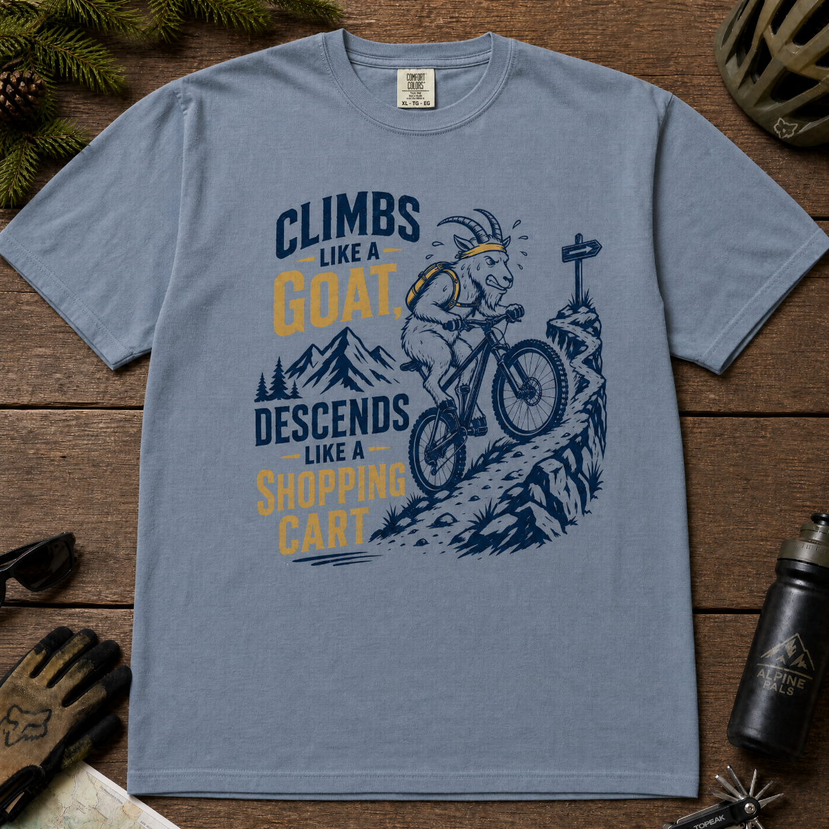 Blue jean t-shirt with graphic design and text on a wooden surface with outdoor gear.