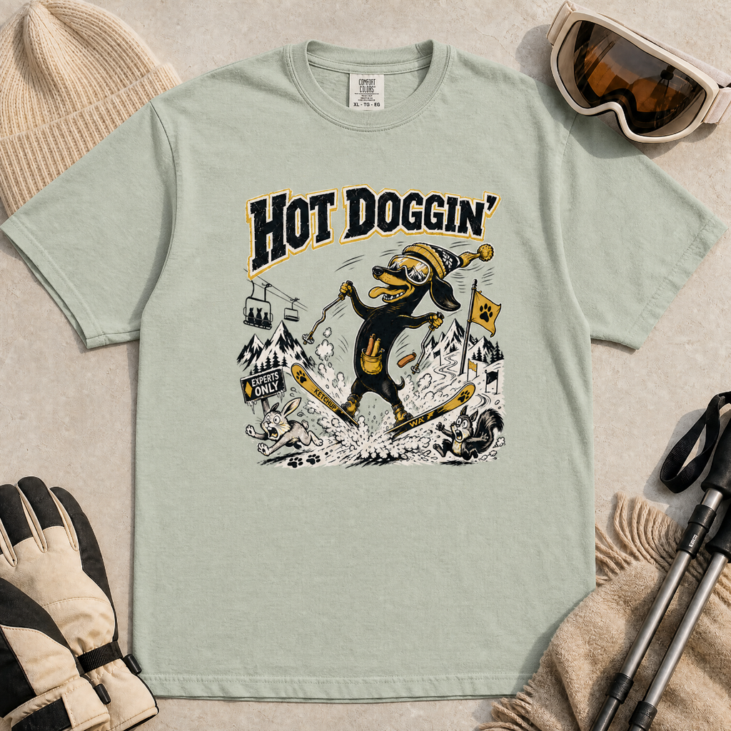 Bay green t-shirt with a graphic design of a dog skiing, text 'Hot Doggin', surrounded by winter accessories on a beige surface.