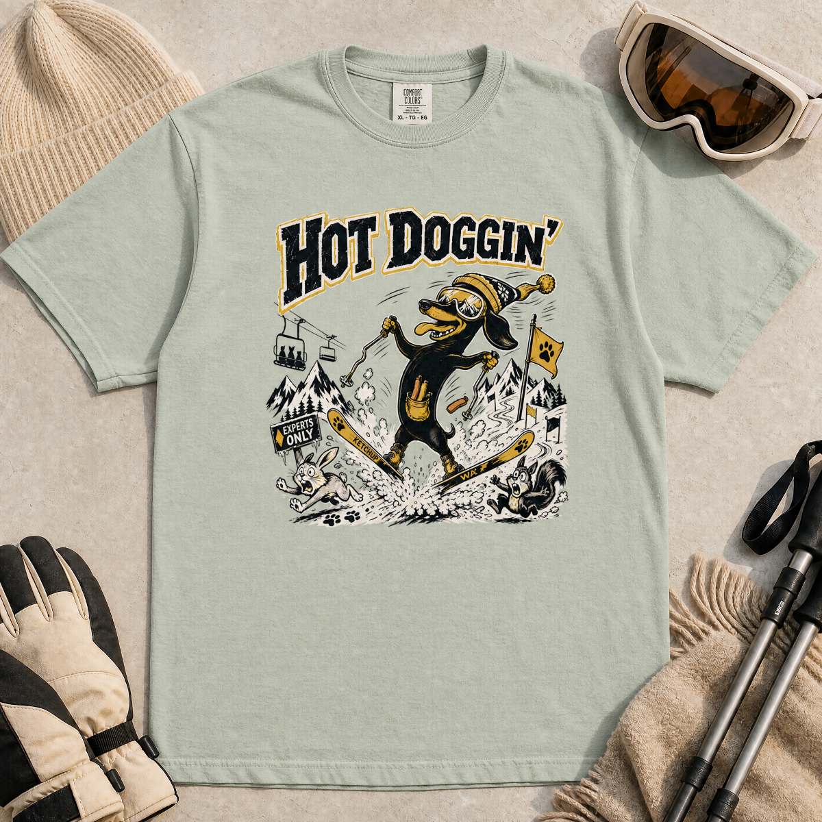 Bay green t-shirt with a graphic design of a dog skiing, text 'Hot Doggin', surrounded by winter accessories on a beige surface.