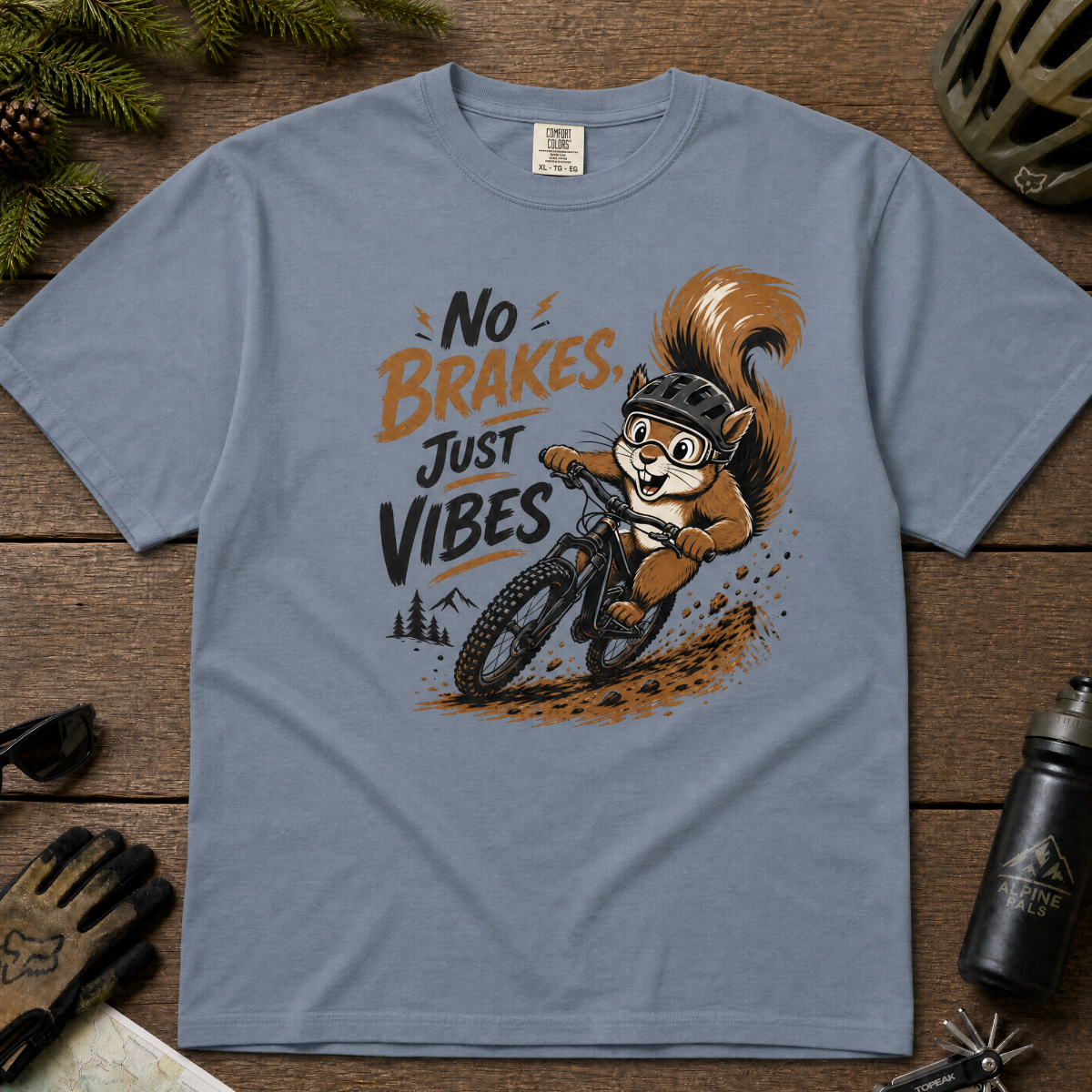Blue jean colored t-shirt with a graphic of a squirrel riding a bike and text 'No Brakes, Just Vibes' on a wooden surface.