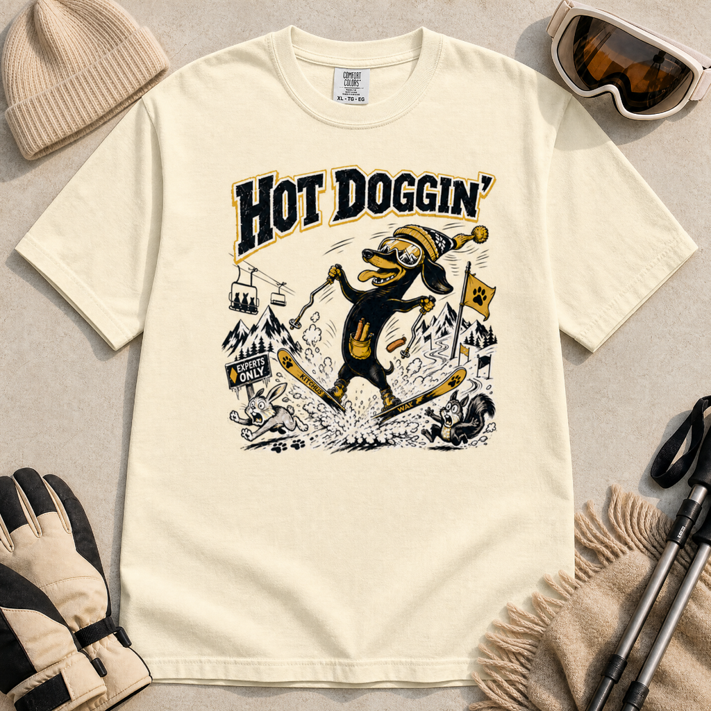 Ivory white t-shirt with a graphic design of a dog skiing, text 'Hot Doggin', surrounded by winter accessories on a beige surface.