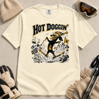 Ivory white t-shirt with a graphic design of a dog skiing, text 'Hot Doggin', surrounded by winter accessories on a beige surface.
