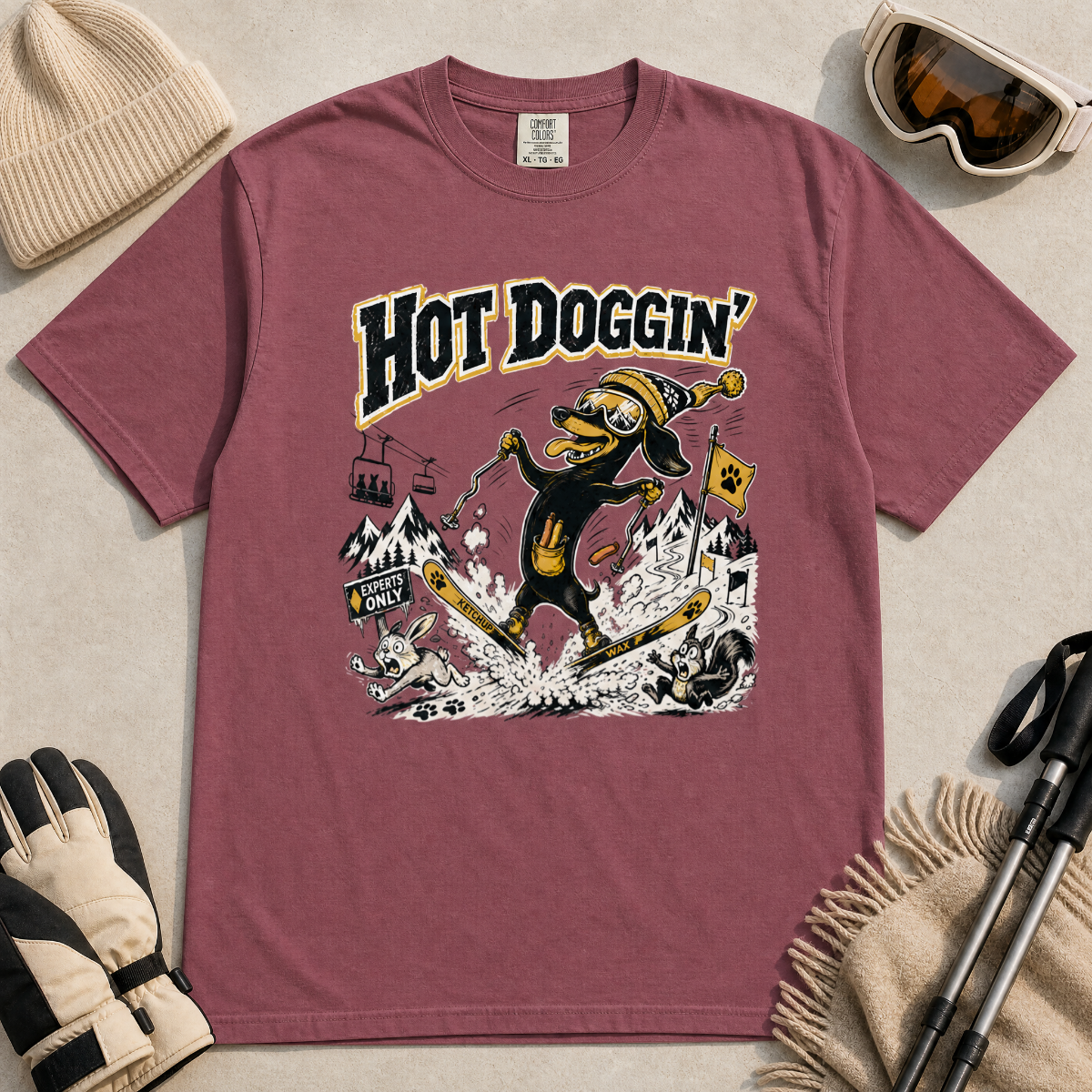 Brick red t-shirt with a graphic design of a dog skiing, text 'Hot Doggin', surrounded by winter accessories on a beige surface.