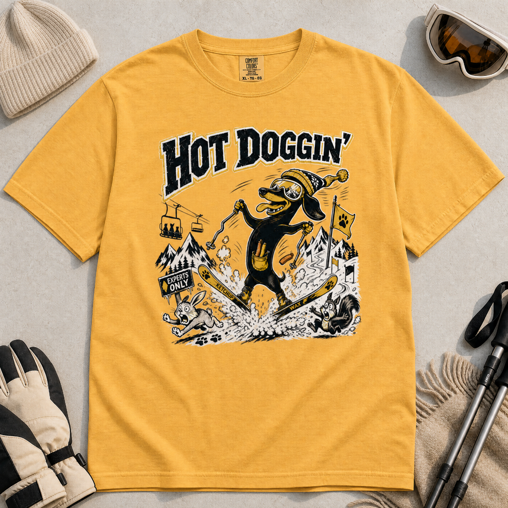 Mustard yellow t-shirt with a graphic design of a dog skiing, text 'Hot Doggin', surrounded by winter accessories on a beige surface.