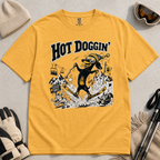 Mustard yellow t-shirt with a graphic design of a dog skiing, text 'Hot Doggin', surrounded by winter accessories on a beige surface.