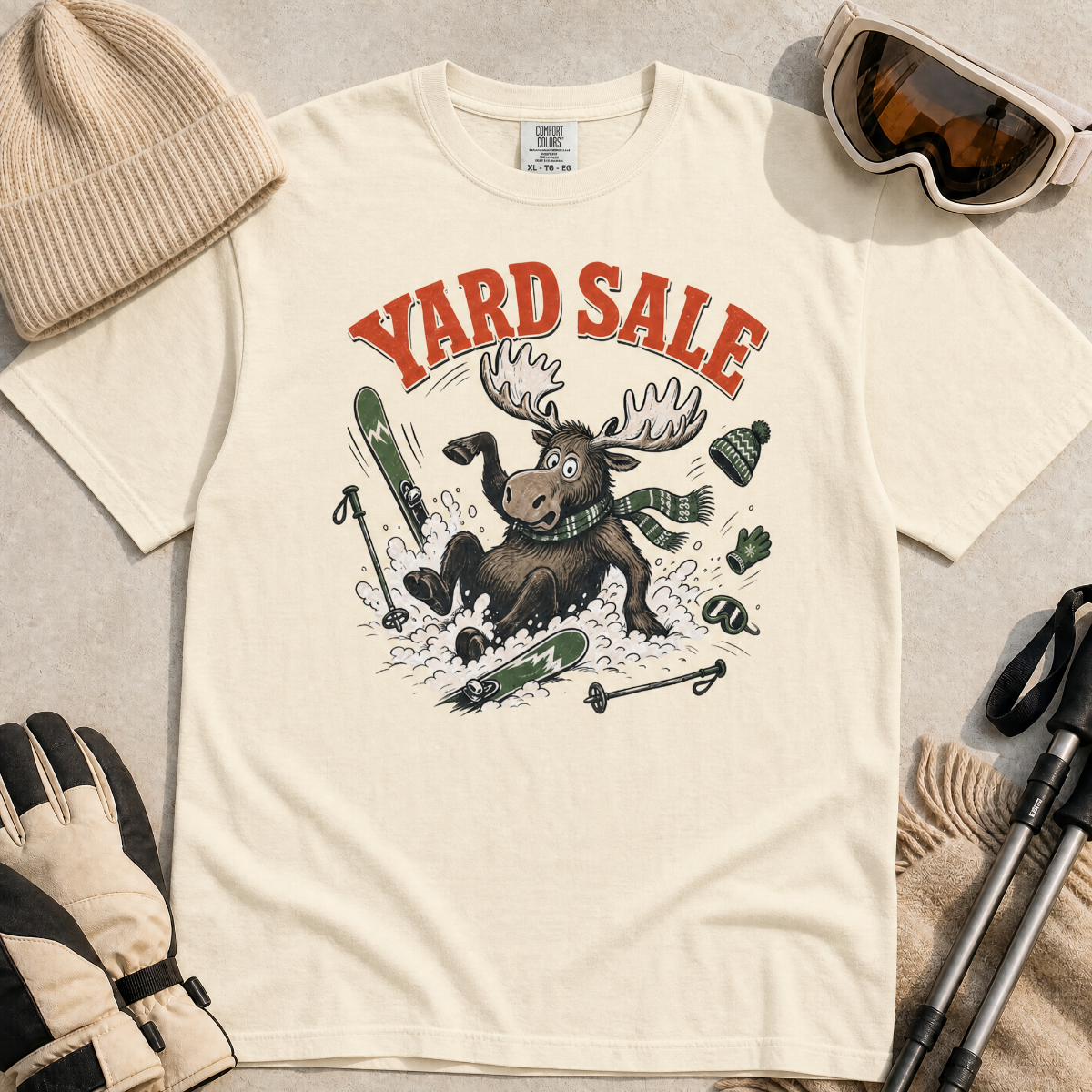 Ivory white t-shirt with moose graphic and 'Yard Sale' text, surrounded by winter accessories on a light background